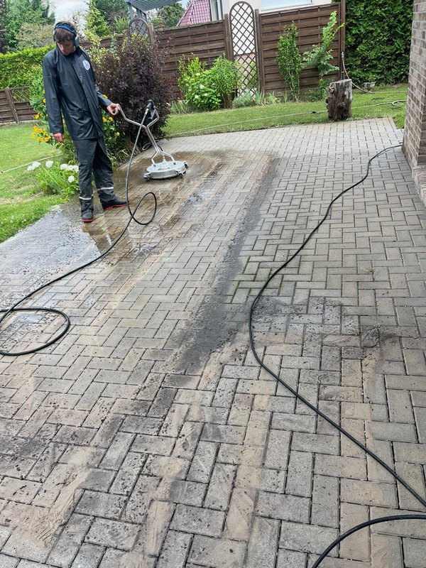 Mitarbeiter führt den Flächenreiniger über eine halb gereinigte Betonpflasterfläche im Garten — echtes Arbeitsfoto